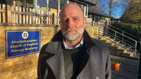 A man in his 50s with a short, grey beard and thinning brown hair stands in front of the entrance of Minchinhampton Church of England Primary Academy. The sign and a set of stairs with railings leading up to the school can be seen behind him. The light is golden as it is the afternoon on a sunny winter day. 