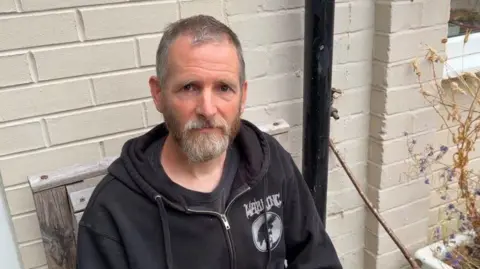 A man with a grey beard and short hair wearing a black hooded top and sitting outside on a wooden chair. 