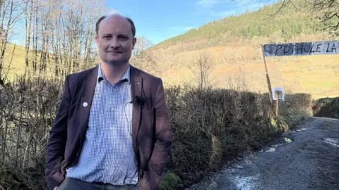 A middle-aged man with receding gray hair stands in front of a sign for Pothole Land on a country lane. He is wearing a brown blazer, blue and white check shirt and faded black jeans. 