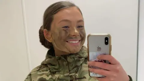 Family Handout A mirror selfie of Gunner Beck wearing her camouflage military uniform, with her hair tied back into a slick low bun. She has camouflage green and brown face paint on and is smiling at the mirror.