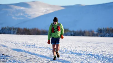 A runner in the Pentland Hills in Edinburgh. He is wearing a black hat and sunglasses and has a white beard. He is wearing a bright green jacket with a red bag along with orange gloves, blue shorts and dark-coloured shoes. Snow is covering the ground and hills behind him.