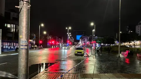 Two police vehicles parked on a road at the scene. It is dark. Street lamps are on. A number of red traffic lights can be seen in the distance. The civic centre building is at the end of the road. 