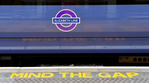 A close-up of a train with a purple "Elizabeth Line" roundel on its side. The platform edge has "MIND THE GAP" written in bold yellow letters. 