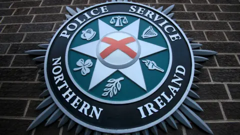 A close-up of the PSNI crest on brown brick wall.  The badge has a red cross in the centre of a star, surrounded by six emblems with include a harp, a crown, a shamrock and the scales of justice.  The centre of the badge is surrounded by a black circle featuring the words Police Service of Northern Ireland in capital letters. 