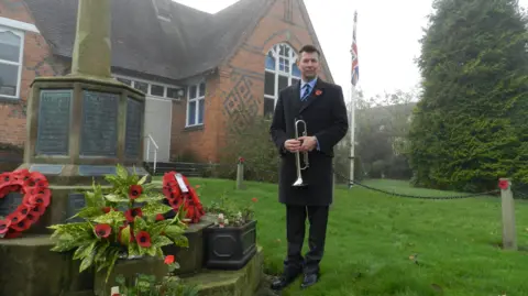 Niall Roberts A man in a dark coat and suit and poppy, holding a bugle, standing beside a memorial with poppies on it. Behind him is a red brick building with some darker bricks in a diamond pattern. A big tree is to the right of the image with a flag pole with the union flag on it, next to it.