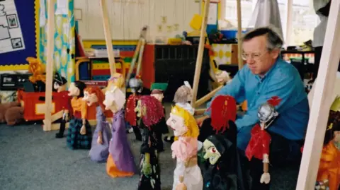 Duschenes family A man crouching down in front of a row of puppets in a school classroom. He has glasses and is wearing a blue shirt.