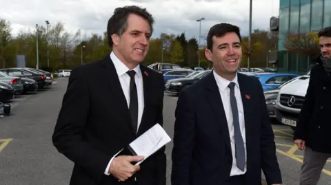 PA Media Two white middle aged men with short hair wearing dark suits, white shirts and ties walk through a car park, smiling broadly. 