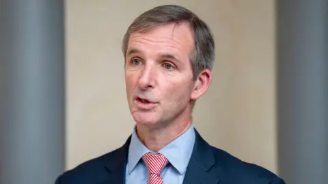 PA Liam McArthur MSP standing against a white and dark blue background. He is wearing a blue suit with a red striped tie. 