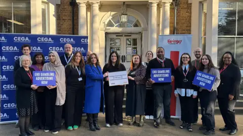 A group of people in front of a big blue board with the words 'CEGA' repeated on the left and on the right, a long, white board saying the words 'Haringey' in red. The group of 16 are a mix of male and female holding posters saying 'End gambling advertising now'.