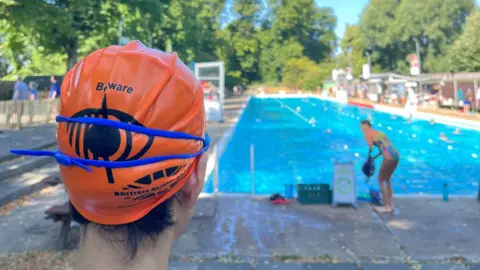 Jean Zhang is facing the lido and away from the camera. She is wearing an orange swimming cap with a large graphic of an eye on the back that has a line through it. It reads 'beware'. Her black hair is coming out from under the cap and onto her neck. Her blue goggles are also over the top of the cap. The lido, in the background, is long and very blue with three lanes marked out. A woman looks to be getting out of the pool. 