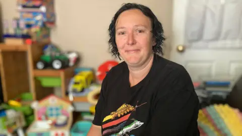 Fran Pedley sits smiling and looking at the camera. She has a black t shirt on, with colourful stripes. In the background are lots of colourful children's toys