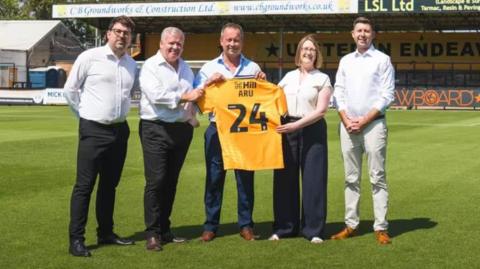 Five people on a football pitch holding a yellow football shirt with the number 24 on it