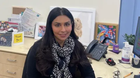 Deepali Misra-Sharp Dr Deepali Misra-Sharp sits at her work desk, with pictures and cards behind her.