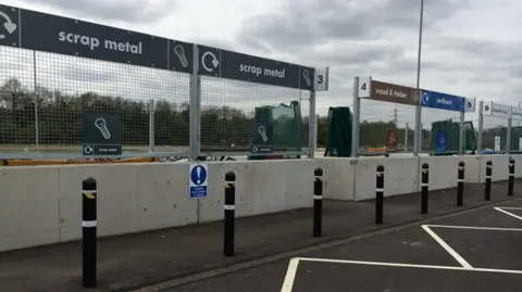 A household waste recycling centre with signs for scrap metal, wood and timber and cardboard. In front of the signs there are concrete barriers and a line of bollards.