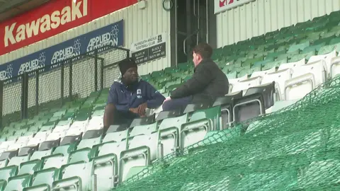 A picture of Brendan Wiredu chatting. He is sat on a green chair wearing a navy coat.