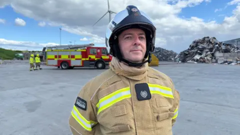 A firefighter in uniform stands at a landfill site with a fire appliance in the background