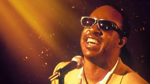 Stevie Wonder as a younger man. He is wearing black sunglasses and has his head back singing with a smile