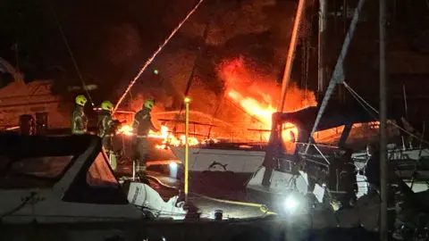 Bright orange flames coming off a boat, with firefighters close to the scene