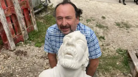 BBC/Jack Hadaway-Weller John David stands behind the grotesque made for him, which will be placed on York Minster. The grotesque is in white.