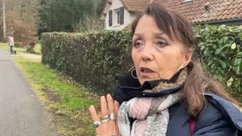 A woman with brunette hair, wearing a coat and scarf, is looking away from the camera. She is stood outside and appears to be mid-conversation