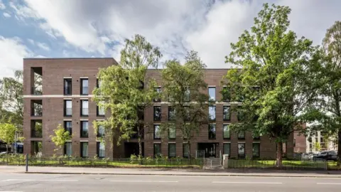 Keith Hunter A brown building with rectangular windows, behind trees, under a cloudy clue sky.