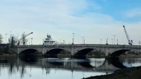 Bridge with oval arches across a river with birds on the water and a ship in the background, against a cloudy blue sky.