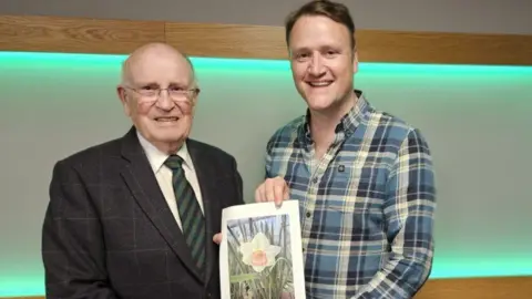 An elderly man in a dark suit and green striped tie stands to the left of another man in a chequered blue shirt. They are both holding a photograph of a daffodil. 