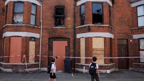 Two red brick terrace houses have smashed upstairs windows. All the windows and doors below have boarded up windows. There is red and white tape running across them both. Two young boys in white shirts and black shorts look towards the houses with their backs to the camera.  