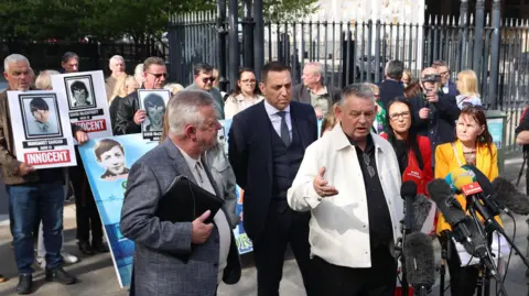 PA Media Harry Gargan speaking in front of microphones. He is wearing a white jacket and black shirt. Two women are two his left, a man wearing a suit is behind him and another man holding a folder is to his right. Other people holding placards of the deceased are standing at the back.