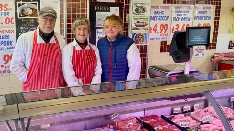 Two women and a man stand behind a glass counter filled with meat. The man on the left is wearing glasses, grey baseball cap, fleece, white butchers coat, red and white apron and has white facial hair. The woman next to him is wearing a butchers coat and apron over a red fleece and has light hair tied back. To the right of her is a woman in a butcher's coat, with a blue bodywarmer over the top and blonde hair tied back. There is a weighing scale till on the counter and posters behind them advertising their meat on a white and maroon tiled wall.