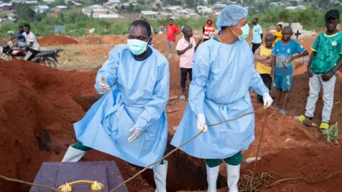 EPA Two people in blue PPE stand over a coffin as they lower it into the ground. Some young boys can be seen in the background looking on.