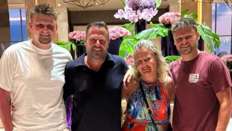 Chenery family Max Chenery (left) has spiky fair hair and a goatee beard. He is wearing a white T-shirt and has his arm around his father Paul, who wearing a navy polo shirt. He has his arm around his wife Tanya, who has blonde wavy hair and is wearing an orange, blue and black halterneck dress. She has her arm around Alfie, who has fair hair and is wearing a burgundy T-shirt. They are all smiling and standing in front of flower arrangements.