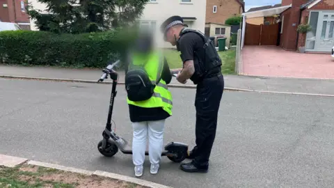 Sam Read/BBC A police officer stands next to a woman seen from behind, next to an e-scooter. She is wearing a high-vis neon vest and grey jogging trousers.