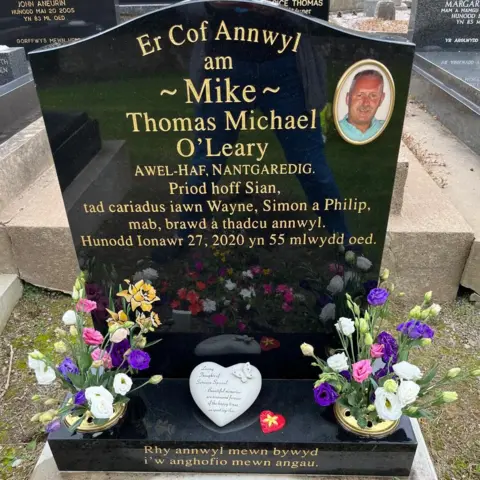 Family photo A black gravestone with a picture of Mike O'Leary, in a golden circle in the top righthand corner. The writing is in Welsh and reads: Er Cof Annwyl am Mike Thomas Michael O'Leary: Awel-Haf, Nantgaredig, Priod hoff Sian, tad cariadus Wayne, Simon a Philip, mab, brawd, a thadcu annwyl. Hunodd Ionawr, 27, 2020 yn 55 mlwydd oed