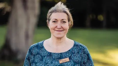 Yorkshire Wildlife Trust A woman with tied-back, light brown hair, smiles at the camera. She is wearing a blue and white patterned dress. 