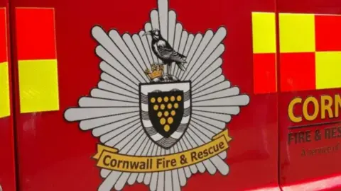 A close-up of the side of a Cornwall Fire and Rescue fire engine. It is red with yellow and red squares. At the centre is the fire service's logo which is grey, has the Cornish badge with a crown and bird on top. Below is a yellow badge with the words "Cornwall Fire & Rescue".