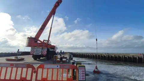 Bridport Harbour / Dorset Council A large orange crane with a grabber is on the quayside scooping up sand from the water below.