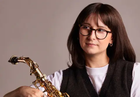 A lady with brown hair and glasses holding a saxophone looking to the left