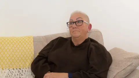 BBC Woman with short hair and glasses sat on a sofa wearing brown jumper 