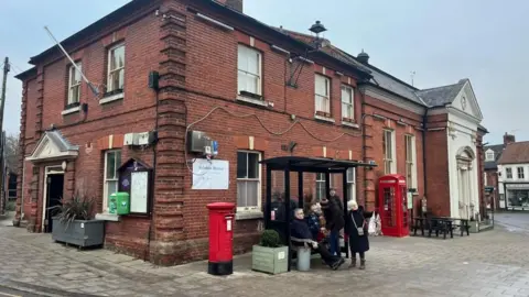 Andrew Turner/BBC Aylsham Town Hall, a red-brick Georgian building with decorated features and a grand entrance to the right of the image. Outside it are a red pillar box, a bus shelter and a red K6 British telephone box.