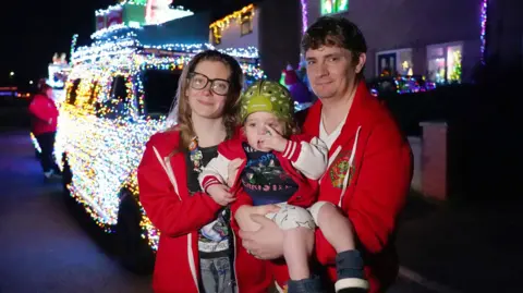Molly and James smile into the camera holding Rory between them. All three are wearing red jackets or jumpers. Behind them a lit up VW van covered in lights can be seen parked next to houses on the street.