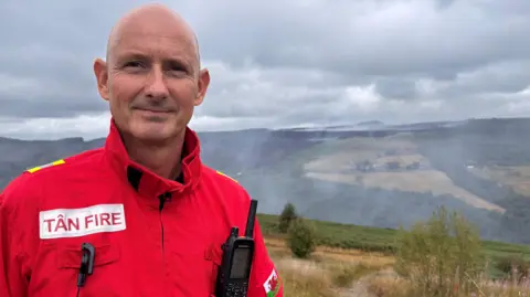 A firefighter in red uniform with a bald head looking at the camera. On his top it says Tan/Fire and he is standing in front of a valley where there is smoke rising.
