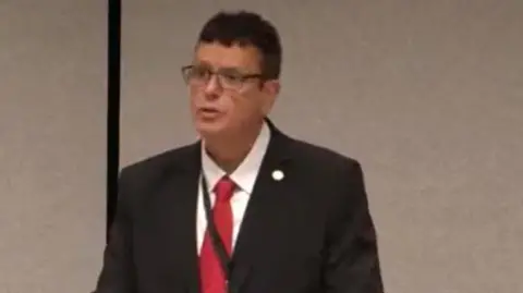 Senedd MS Nigel Williams in the Senedd. He has short dark hair and glasses and wears a black suit jacket, white shirt with a red tie. 