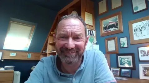 David Blevings is a middle aged man with short, greying brown hair and a beard. He is wearing a blue polo-shirt and is smiling at the camera. Behind him is an office with the walls painted blue and lots of pictures on on them. 