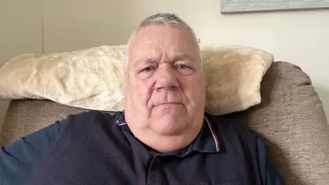 Kevin Newton sat on a chair looking directly into the camera. He is wearing a blue top.