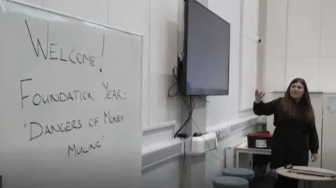 Criminology lecturer Dr Sam Mapston stood in a class room. She is pointing at a screen. Next to the screen is a white board with the words 'Welcome Foundation Year: Dangers of Money Muling' written on it. 