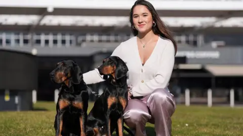 PA Media A woman with a white buttoned top and mauve jeans squats on a grass field next to two dogs. The dogs have a mix of black and brown fur and look to the left. The woman has long dark brown hair, past her shoulders.