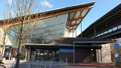 Glass building with sloped roof. A smaller reception structure in-front and to the side. There are trees and bollards with paving.  