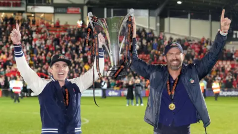 PA Media Ryan Reynolds and Rob McElhenney holding the National League Trophy on the pitch