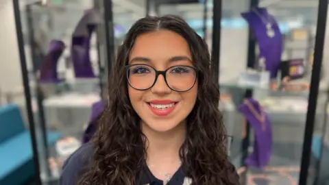 A woman looks into the camera smiling with red lipstick and long dark hair past her shoulders. She wears wide-rimmed, black glasses.